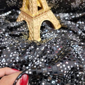 May include: A gold Eiffel Tower ornament sits on black fabric with square, silver reflective details. The fabric is gathered, creating folds and shadows. The image is well-lit, highlighting the textures.