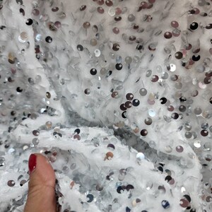 May include: Close-up of a white fabric covered in silver sequins. The sequins are round and scattered across the surface, reflecting light. The fabric appears soft and textured, creating a shimmering effect.