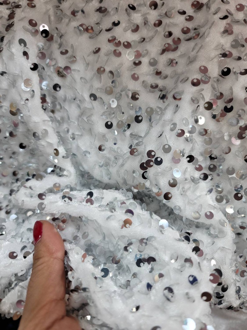May include: Close-up of a white fabric covered in silver sequins. The sequins are round and scattered across the surface, reflecting light. The fabric appears soft and textured, creating a shimmering effect.