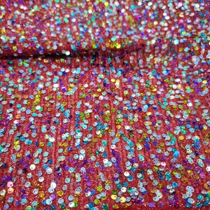 May include: Close-up of red fabric covered in iridescent sequins. The sequins are small and round, reflecting a rainbow of colors. The fabric appears to be slightly textured, with a subtle sheen. The overall effect is sparkly and festive.