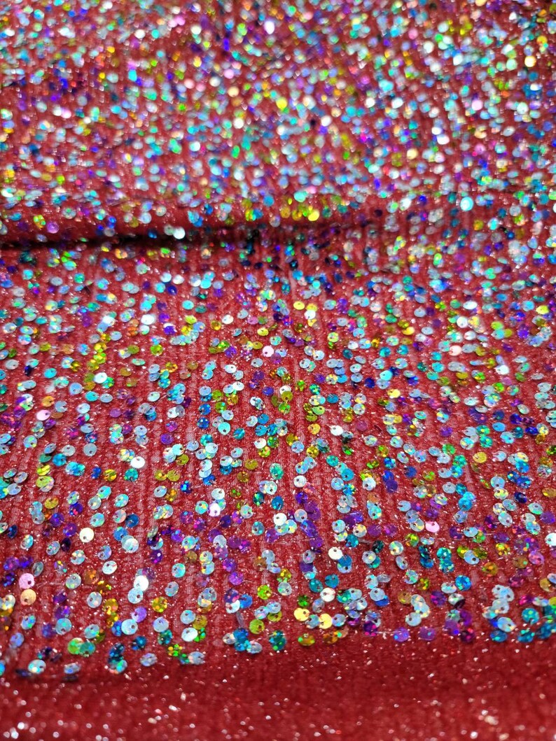 May include: Close-up of red fabric covered in iridescent sequins. The sequins are small and round, reflecting a rainbow of colors. The fabric appears to be slightly textured, with a subtle sheen. The overall effect is sparkly and festive.