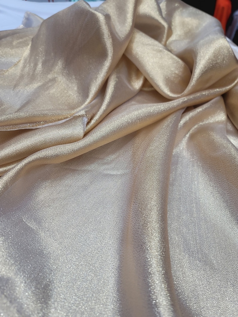 May include: A close-up of a gold satin fabric with a subtle shimmer. The fabric is draped and folded, showcasing its soft texture.