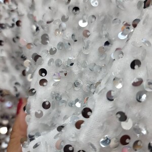 May include: Close-up of a white fabric covered in silver sequins. The sequins are round and reflective, catching the light. The fabric appears soft and textured, creating a shimmering effect.