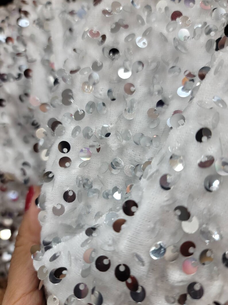 May include: Close-up of a white fabric covered in silver sequins. The sequins are round and reflective, catching the light. The fabric appears soft and textured, creating a shimmering effect.