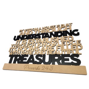 May include: A wooden wall decor with the text "BY WISDOM A HOUSE IS BUILT AND THROUGH UNDERSTANDING IT IS ESTABLISHED THROUGH KNOWLEDGE ITS ROOMS ARE FILLED WITH RARE AND BEAUTIFUL TREASURES Proverbs 24:3" in black and brown letters.