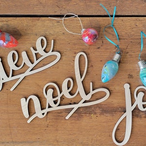 May include: Wooden cursive script cutouts spelling "believe", "noel", and "joy", with painted Christmas light bulb ornaments. The bulbs are painted in red, blue, and green, with string for hanging. The items are on a wooden surface.