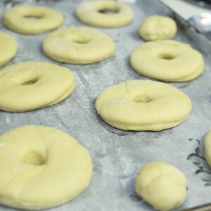 May include: A close-up shot of uncooked dough for donuts on a baking sheet lined with parchment paper. The dough is a pale yellow color, with a round shape and a hole in the center. Several round dough balls are also visible.