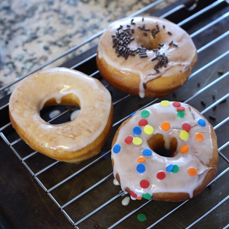 May include: Three glazed donuts on a metal cooling rack. One donut has chocolate sprinkles, another has colorful sprinkles, and the third is plain. The donuts have a light brown color and a glossy glaze.