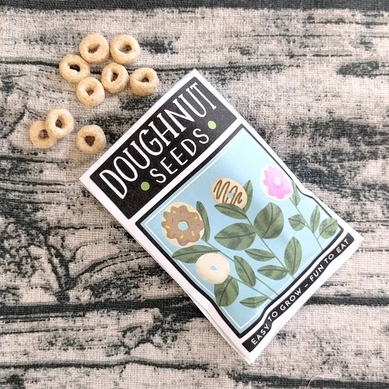 May include: A packet of "DOUGHNUT SEEDS" with a light blue background featuring illustrated donuts. The packet is next to a small pile of donut-shaped cereal pieces. Text on the packet reads "EASY TO GROW - FUN TO EAT."