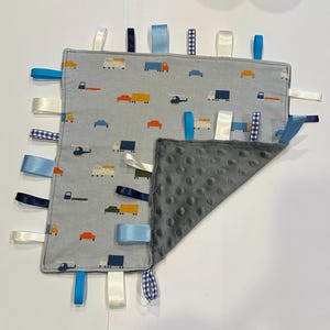 May include: A gray baby tag blanket with a transportation pattern featuring cars, trucks, and helicopters. The blanket has a soft, gray, dimpled backing and colorful satin ribbons in various colors and patterns around the edges.