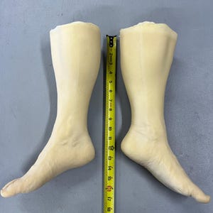 May include: A pair of pale, skin-toned mannequin legs and feet positioned side-by-side. A yellow measuring tape is placed vertically between them, showing a total length of approximately 20 inches from the top of the calf to the tip of the toes.