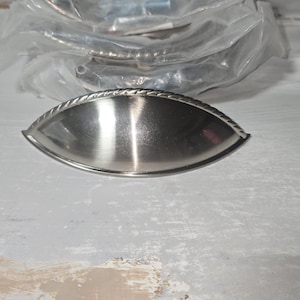 May include: A silver-toned metal cup pull handle featuring a smooth, arched center and a decorative rope-style border along the top edge. The hardware has a brushed finish and a semi-circular shape, designed for attachment to drawers or cabinets.