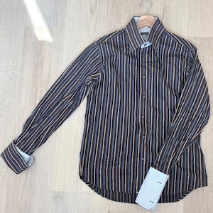 May include: A long-sleeved button-down shirt featuring a vertical striped pattern in dark brown, light blue, and tan. The shirt has a structured collar, a front button placket, and light blue French cuffs that are folded back to reveal the interior.