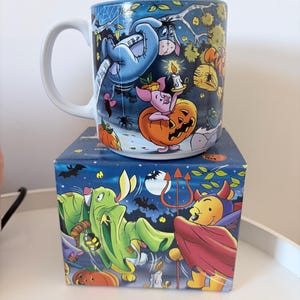 May include: A white ceramic mug with a Halloween-themed design featuring Winnie the Pooh characters. The mug is on a matching box. The design includes Eeyore, Piglet, and Pooh Bear in Halloween costumes, with pumpkins and bats.