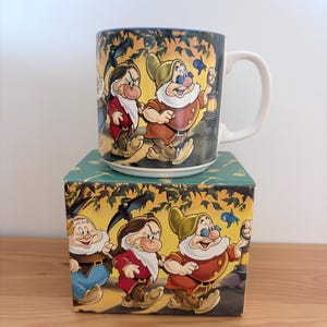 Könnte beinhalten: Ein Keramikbecher mit weißem Henkel und einer umlaufenden Illustration von drei Zeichentrickzwergen, die durch einen Wald gehen. Das Design zeigt Figuren mit weißen Bärten, bunten Tuniken und einer Laterne, passend zum quadratischen Karton.