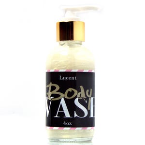 May include: A clear glass bottle of body wash with a white pump and gold cap. The black label reads "Lucent Body Wash" in gold and white lettering. The bottle contains a shimmering, light-colored liquid and is 4 oz.