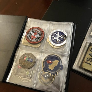 May include: A collection of military challenge coins and patches displayed in a black album. The coins feature various designs, including military unit insignias, flags, and text. The album pages are clear plastic, allowing visibility of the coins.