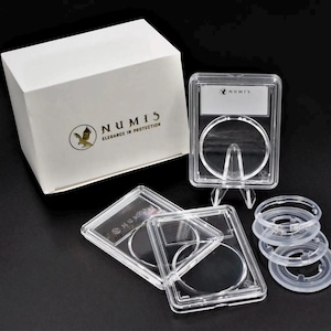 May include: A white box with the text "NUMIS Elegance in Protection" and a logo, along with several clear plastic coin holders and rings. The holders are rectangular with a circular opening, designed to display and protect coins.