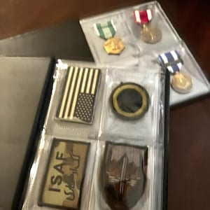 May include: A black album with clear sleeves displaying military medals and patches. The patches include an American flag design, an ISAF patch, and a shield-shaped emblem. The medals have red, white, blue, and green ribbons.