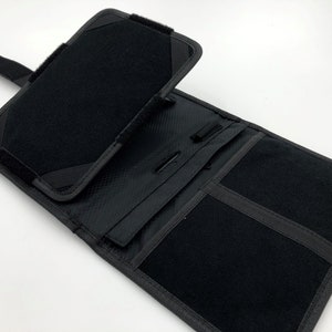 May include: Black tablet case with multiple compartments and a Velcro closure. The case is made of a textured material and features a flap that folds over to secure the contents. The interior includes pockets and a pen holder.