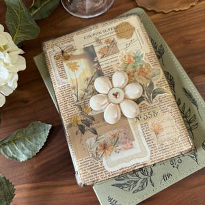 Peut inclure: Un journal de style vintage avec un motif floral et une fleur blanche avec un bouton doré au centre. Le journal est ouvert et a une couverture verte avec un motif de feuilles. Le texte "COUPON SUPERIEUR QUALITE EXT" est visible sur le journal.