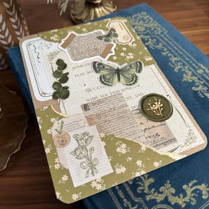 Könnte beinhalten: Ein grünes und braunes Journal mit einem Blumenmuster und mehreren dekorativen Aufklebern. Die Aufkleber beinhalten einen Schmetterling, ein Wachssiegel und ein Blumenmuster. Der Text auf dem Journal lautet "Some of flat. So... But even while you find someone..."