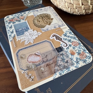 May include: A decorative journal with a collage design featuring seashells, a seahorse, and floral patterns. The journal has a blue cover with gold accents and a textured, beige center. The collage includes stamps and handwritten script.