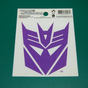 Könnte beinhalten: Lilafarbener Transformers-Logo-Aufkleber mit dem Text "Transformers" und "www.palisadetoys.com".