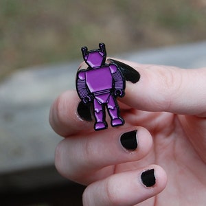 Puede incluir: Un pin de esmalte de robot púrpura con contornos negros. El pin presenta un diseño de robot estilizado con un cuerpo y antenas detallados. El pin es de color púrpura vibrante y es probablemente un artículo de colección o un accesorio.
