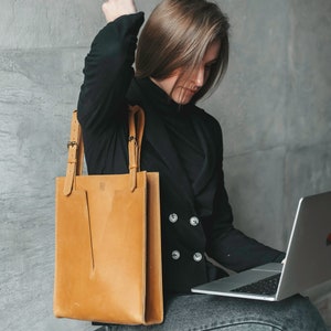 A woman sitting on a chair with the laptop. She is looking on the screen of the laptop. She is holding a yellow leather tote on her right shoulder.