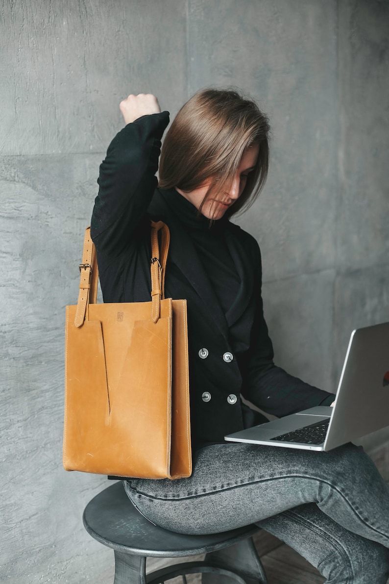 A woman sitting on a chair with the laptop. She is looking on the screen of the laptop. She is holding a yellow leather tote on her right shoulder.