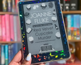Puede incluir: Un lector electrónico con una funda protectora de acabado oscuro, iridiscente y con purpurina holográfica. La pantalla muestra la portada del libro Red Velvet Cupcake Murder de Joanne Fluke, con ilustraciones de cupcakes, una botella y un edificio.
