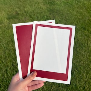 May include: Two blank red photo frame stickers with white borders. The stickers are held in a hand against a green grassy background.