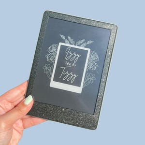 May include: A black e-reader with a glittery silver border is held against a light blue background. The screen displays the text "Izzy in a Tizzy" inside a white frame, surrounded by floral line art.