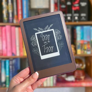 May include: A bronze-colored e-reader featuring a white rectangular sticker on the screen. The sticker displays a line-art illustration of roses and wheat surrounding the white text "Izzy in a Tizzy" written in a cursive script font.