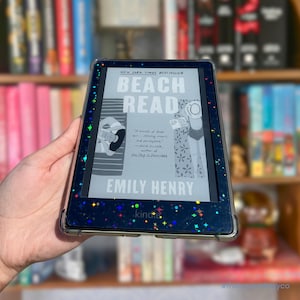 May include: A black e-reader device featuring a clear protective case with a holographic star pattern. The screen displays the book cover for Beach Read by Emily Henry, showing a graphic illustration of two people on beach towels under the sun.