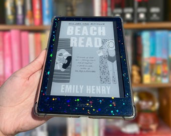 Könnte beinhalten: Ein schwarzer E-Reader mit einer transparenten Schutzhülle, die mit einem holografischen Sternenmuster versehen ist. Auf dem Bildschirm ist das Buchcover von Beach Read von Emily Henry mit einer Grafik von zwei Personen am Strand zu sehen.