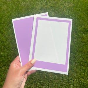 May include: Two purple blank photo frame stickers with a white border. The stickers are on a green grassy background.