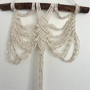 May include: Cream-colored macrame wall hanging. The design features a central diamond pattern with cascading knotted strands, suspended from a natural wooden branch. The piece is a decorative, bohemian-style home decor item.