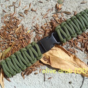 May include: A green paracord bracelet with a black plastic buckle. The bracelet is on a light brown surface with brown leaves.