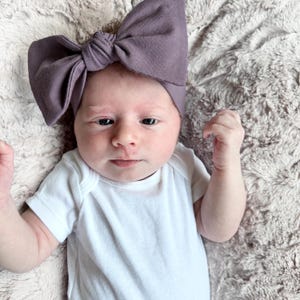 May include: A baby wearing a white onesie with a large purple bow headband. The baby is lying on a light brown fluffy blanket.