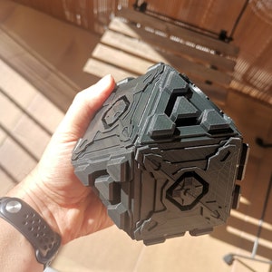 Puede incluir: Un objeto negro en forma de cubo con una superficie texturizada y detalles intrincados. El objeto tiene un diseño geométrico con múltiples paneles y parece estar hecho de plástico.