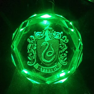 Puede incluir: Un pisapapeles de vidrio verde con un grabado detallado del escudo de la casa de Hogwarts para Slytherin. El escudo presenta una serpiente y las palabras "Slytherin" en una pancarta.