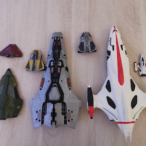 Puede incluir: Una colecci&oacute;n de seis naves espaciales en miniatura, que incluyen una gran nave espacial gris y plateada, una nave espacial blanca con detalles en negro y rojo, una nave espacial verde, una nave espacial amarilla y dos naves espaciales grises m&aacute;s peque&ntilde;as.