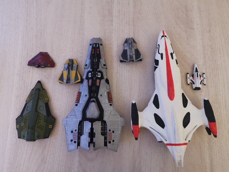 Puede incluir: Una colecci&oacute;n de seis naves espaciales en miniatura, que incluyen una gran nave espacial gris y plateada, una nave espacial blanca con detalles en negro y rojo, una nave espacial verde, una nave espacial amarilla y dos naves espaciales grises m&aacute;s peque&ntilde;as.