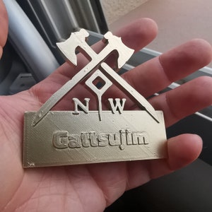 May include: A gold-colored metal sign with two crossed axes and the letters "NW" above the word "GattsuJim".