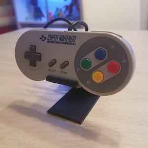 Op de afbeelding: Een grijze Super Nintendo Entertainment System-controller met een zwarte standaard. De controller heeft een zwarte D-pad, vier gekleurde knoppen en een start- en een select-knop.