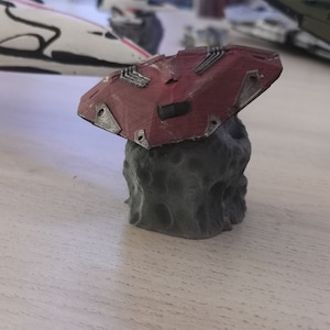 Puede incluir: Un modelo de nave espacial impreso en 3D de color rojo y gris que descansa sobre una roca. La nave espacial tiene un dise&ntilde;o detallado con m&uacute;ltiples paneles y una superficie texturizada.
