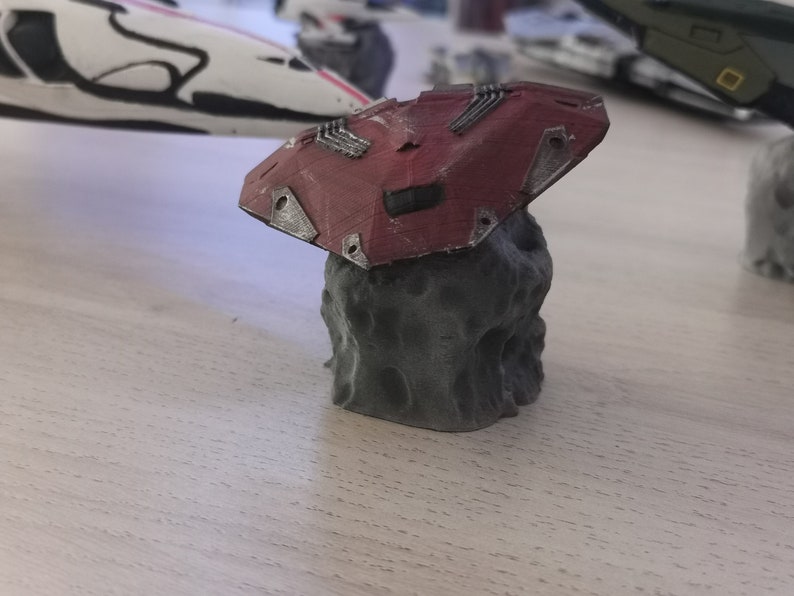 Puede incluir: Un modelo de nave espacial impreso en 3D de color rojo y gris que descansa sobre una roca. La nave espacial tiene un dise&ntilde;o detallado con m&uacute;ltiples paneles y una superficie texturizada.