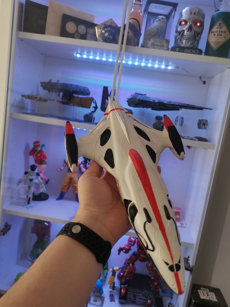 Puede incluir: Un modelo de nave espacial blanco y rojo con detalles negros. El modelo se sostiene en la mano de una persona.